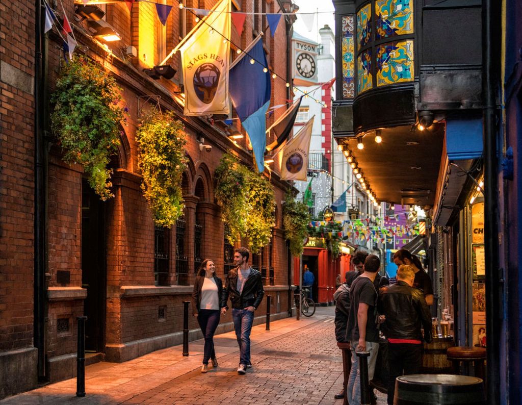 Een levendig, smal straatbeeld in Dublin bij schemering met groepen mensen die wandelen en kletsen. Bakstenen gebouwen versierd met hangende planten en kleurrijke banieren langs de straat. Warme lichten verlichten het geplaveide pad, wat een gezellige sfeer creëert onder een zacht gloeiende lucht - perfect voor een rondreis Ierland. | Echt Ierland Vakanties