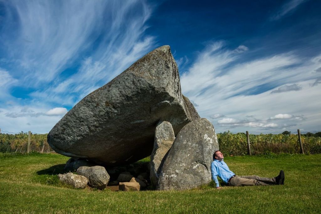 Een man in een blauw shirt en bruine broek zit op gras, leunend tegen een grote, onregelmatig gevormde stenen constructie. De lucht is voornamelijk blauw met strepen wolken. Dit pittoreske tafereel van een serene omgeving is perfect voor degenen die hun vakantie in Ierland koesteren of dromen van een ideale vakantie in Ierland. | Echt Ierland Vakanties
