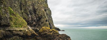 Ervaar het adembenemende uitzicht op de kustklif tijdens uw vakantie in Ierland, met ruige, met mos bedekte rotsen die naar de serene zee leiden. Een schilderachtige houten brug siert de klif. De groenige tint van de oceaan contrasteert met de dikke grijze wolken erboven, terwijl zeevogels fladderen bij de steile rotswand. | Echt Ierland Vakanties