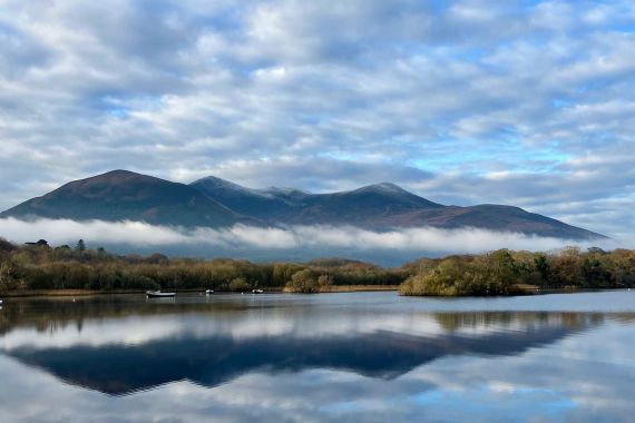 Een sereen meer in Ierland weerspiegelt een bewolkte lucht en verre bergen. Lage mist drijft over de voet van de heuvels. Verschillende kleine boten liggen aangemeerd langs de waterkant, terwijl kalme, reflecterende wateren worden omlijst door weelderig groen, wat u uitnodigt voor een vredige urlaub Ierland. | Echt Ierland Vakanties