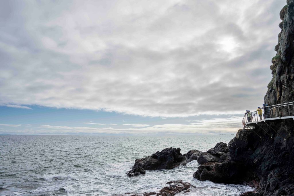 Een dramatisch zeegezicht langs de ruige kust van Ierland ontvouwt zich onder een uitgestrekte lucht. De horizon ontmoet de woelige zee. Rechts kijken mensen op een metalen loopbrug die aan een klif is bevestigd, onderdeel van veel vakanties in Ierland, uit over de oceaan terwijl er door de bewolkte lucht een vleugje blauw te zien is. | Echt Ierland Vakanties