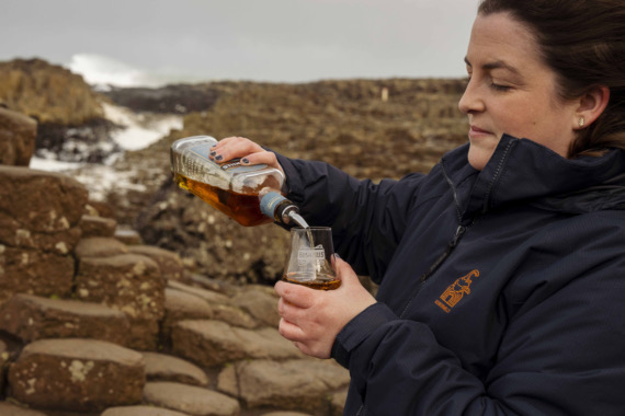 Een persoon in een marineblauw jasje giet whisky uit een fles in een glas met het opschrift "Bushmills". Ze staan op de iconische rotsformatie van Ierland met zeshoekige stenen, die de essentie van een vakantie in Ierland vastleggen. Op de achtergrond piepen witte kliffen door een bewolkte lucht. | Echt Ierland Vakanties