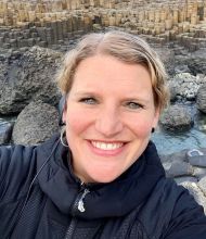Een persoon met een grote glimlach neemt een selfie op de Giant's Causeway, een iconische plek die perfect is voor mensen die op vakantie zijn in Ierland. Ze dragen een zwart jasje en hebben licht haar naar achteren gekamd. De achtergrond toont de unieke zeshoekige basaltkolommen en een vleugje water. | Echt Ierland Vakanties