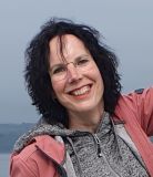 Lachende vrouw met krullend zwart haar en een bril, gekleed in een grijze hoodie met patroon en roze jas, staat buiten tegen een wazige, bewolkte lucht en een ver landschap. Haar vrolijke uitdrukking en casual kleding hinten naar de vreugde van een "vakantie Ierland" te midden van het serene landschap. | Echt Ierland Vakanties