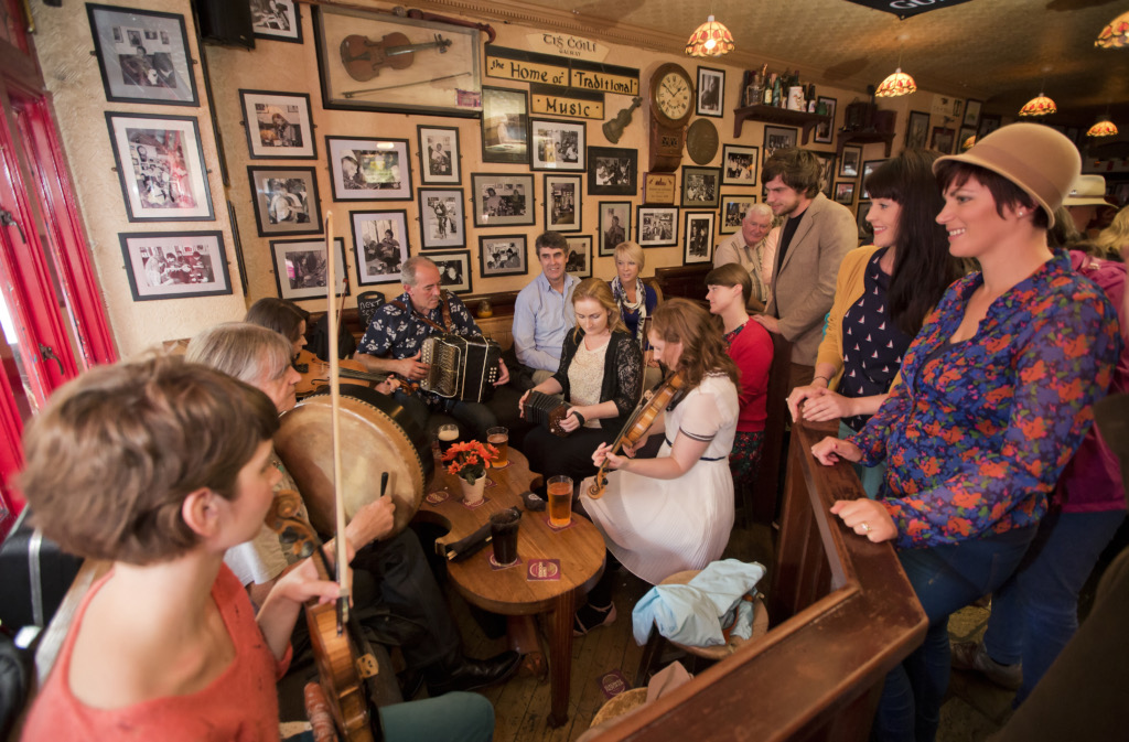Een levendige groep muzikanten bespeelt traditionele instrumenten als viool en accordeon in een gezellige pub, een perfect momentopname van een vakantie in Ierland. Omringd door lachende toeschouwers en ingelijste foto's aan de muren, staan drankjes op houten tafels, wat een warme, bruisende sfeer creëert. | Echt Ierland Vakanties