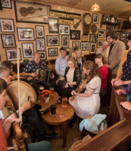 Een levendige groep muzikanten bespeelt traditionele instrumenten als viool en accordeon in een gezellige pub, een perfect momentopname van een vakantie in Ierland. Omringd door lachende toeschouwers en ingelijste foto's aan de muren, staan drankjes op houten tafels, wat een warme, bruisende sfeer creëert. | Echt Ierland Vakanties
