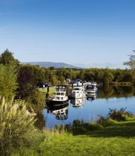 Een serene rivieroever die doet denken aan een idyllische rondreis Ierland, met boten afgemeerd langs een kalme, met bomen omzoomde rivier. Weelderig groen omringt het gebied, hoog gras op de voorgrond en verschillende bomen op de achtergrond. Een helderblauwe lucht boven ons hoofd complementeert de verre heuvels aan de horizon. | Echt Ierland Vakanties