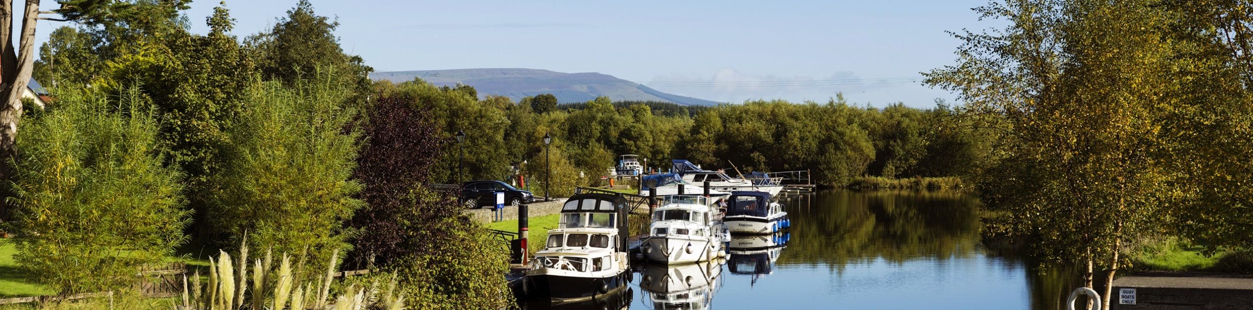 Een serene rivieroever die doet denken aan een idyllische rondreis Ierland, met boten afgemeerd langs een kalme, met bomen omzoomde rivier. Weelderig groen omringt het gebied, hoog gras op de voorgrond en verschillende bomen op de achtergrond. Een helderblauwe lucht boven ons hoofd complementeert de verre heuvels aan de horizon. | Echt Ierland Vakanties