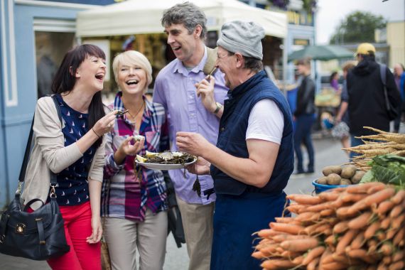 Een groep van drie mensen staat buiten in Ierland en proeft vrolijk het eten dat een verkoper met hen deelt. Ze staan bij een marktkraam met verse wortels en aardappelen en genieten van hun reisen Irland-avontuur, omringd door andere kraampjes en mensen op een zonnige dag. | Echt Ierland Vakanties