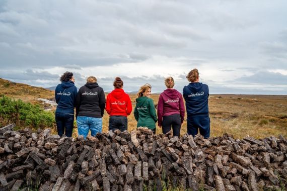 Zes mensen staan met hun rug naar de camera, kijkend naar een uitgestrekt, bewolkt landschap met gras en rotsen tijdens hun rondreis Ierland. Ze dragen kleurrijke hoodies met tekst op de achterkant en zitten op een stapel turf. De bewolkte lucht vormt een dramatische achtergrond voor de aardse tinten van dit Ierse tafereel. | Echt Ierland Vakanties