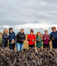 Zeven mensen, vier vrouwen en drie mannen, staan buiten bij een grote stapel turfstenen op een bewolkte dag tijdens hun rundreise Irland. Ze zijn casual gekleed in jasjes en jeans en houden elk een steen vast tegen de achtergrond van een graslandschap en een bewolkte lucht. Ze lijken vrolijk en betrokken. | Echt Ierland Vakanties