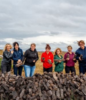 Zeven mensen, vier vrouwen en drie mannen, staan buiten bij een grote stapel turfstenen op een bewolkte dag tijdens hun rundreise Irland. Ze zijn casual gekleed in jasjes en jeans en houden elk een steen vast tegen de achtergrond van een graslandschap en een bewolkte lucht. Ze lijken vrolijk en betrokken. | Echt Ierland Vakanties