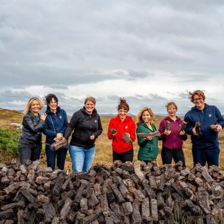 Zeven mensen, vier vrouwen en drie mannen, staan buiten bij een grote stapel turfstenen op een bewolkte dag tijdens hun rundreise Irland. Ze zijn casual gekleed in jasjes en jeans en houden elk een steen vast tegen de achtergrond van een graslandschap en een bewolkte lucht. Ze lijken vrolijk en betrokken. | Echt Ierland Vakanties