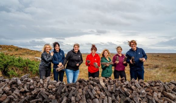 Zeven mensen, vier vrouwen en drie mannen, staan buiten bij een grote stapel turfstenen op een bewolkte dag tijdens hun rundreise Irland. Ze zijn casual gekleed in jasjes en jeans en houden elk een steen vast tegen de achtergrond van een graslandschap en een bewolkte lucht. Ze lijken vrolijk en betrokken. | Echt Ierland Vakanties