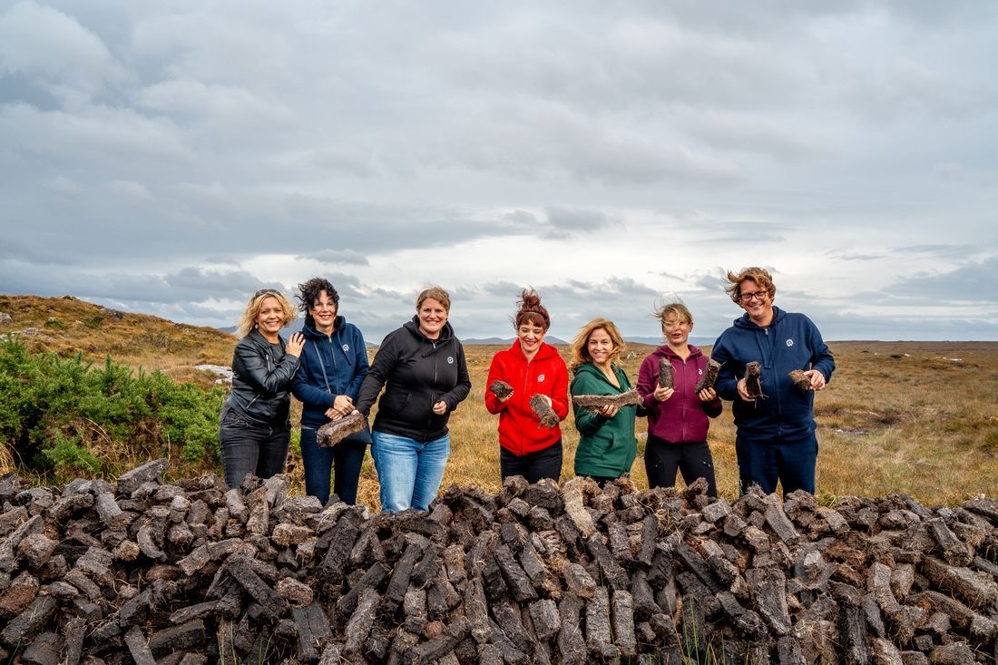 Zeven mensen, vier vrouwen en drie mannen, staan buiten bij een grote stapel turfstenen op een bewolkte dag tijdens hun rundreise Irland. Ze zijn casual gekleed in jasjes en jeans en houden elk een steen vast tegen de achtergrond van een graslandschap en een bewolkte lucht. Ze lijken vrolijk en betrokken. | Echt Ierland Vakanties