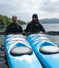 Twee mensen in bijpassende zwarte jassen zitten in blauwe kajaks op een kalm meer in Ierland, peddels vasthoudend en lachend. Het water weerspiegelt de bewolkte lucht. Bomen staan aan één kant van het meer, met bergen zichtbaar op de achtergrond - een perfect moment van hun rondreis Ierland. | Echt Ierland Vakanties