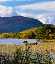 Een rustig meer in Ierland toont twee kleine boten op kalm water, omringd door hoog riet. Weelderige groene en gele bomen langs de oever, die de herfsttinten weerspiegelen. Op de achtergrond voegt een berg onder een gedeeltelijk bewolkte lucht diepte toe aan het serene landschap - een perfecte plek voor een urlaub Ierland. | Echt Ierland Vakanties