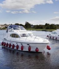 Twee witte motorboten met rode spatborden varen over een brede rivier onder een blauwe lucht met wolken, wat de serene vergezichten weerspiegelt die u tijdens een vakantie in Ierland kunt aantreffen. De leidende boot heeft een overdekt bovendek. Rivieroevers omzoomd met bomen en een verre brug maken het idyllische tafereel compleet, bezaaid met verschillende boten op de achtergrond. | Echt Ierland Vakanties