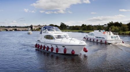 Twee witte motorboten met rode spatborden varen over een brede rivier onder een blauwe lucht met wolken, wat de serene vergezichten weerspiegelt die u tijdens een vakantie in Ierland kunt aantreffen. De leidende boot heeft een overdekt bovendek. Rivieroevers omzoomd met bomen en een verre brug maken het idyllische tafereel compleet, bezaaid met verschillende boten op de achtergrond. | Echt Ierland Vakanties