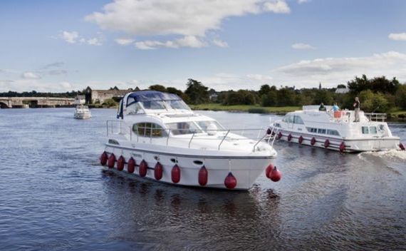 Twee witte motorboten met rode spatborden varen over een brede rivier onder een blauwe lucht met wolken, wat de serene vergezichten weerspiegelt die u tijdens een vakantie in Ierland kunt aantreffen. De leidende boot heeft een overdekt bovendek. Rivieroevers omzoomd met bomen en een verre brug maken het idyllische tafereel compleet, bezaaid met verschillende boten op de achtergrond. | Echt Ierland Vakanties