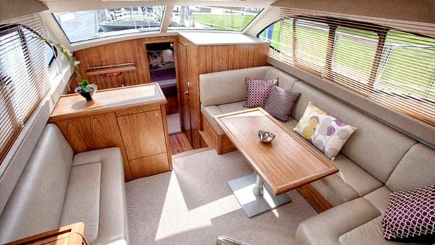 Het interieur van de hut op deze dromerige "vakantie Ierland"-ontsnapping beschikt over L-vormige beige banken versierd met decoratieve kussens rond een houten tafel. Een kleine houten kast met een potplant staat tegen de muur, terwijl grote ramen met jaloezieën natuurlijk licht binnenlaten en de gezellige vloerbedekking verlichten. | Echt Ierland Vakanties