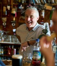Een oudere man, glimlachend en gekleed in een licht shirt, staat achter een bar in een levendige pub in Dublin. Op de voorgrond nipt een vrouw rechts van haar drankje met een rietje, terwijl een ander persoon links wazig is. Flessen en barartikelen vullen de achtergrond en vangen de warme essentie van een onvergetelijke vakantie in Ierland. | Echt Ierland Vakanties
