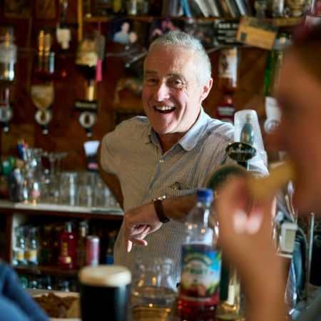 Een oudere man, glimlachend en gekleed in een licht shirt, staat achter een bar in een levendige pub in Dublin. Op de voorgrond nipt een vrouw rechts van haar drankje met een rietje, terwijl een ander persoon links wazig is. Flessen en barartikelen vullen de achtergrond en vangen de warme essentie van een onvergetelijke vakantie in Ierland. | Echt Ierland Vakanties