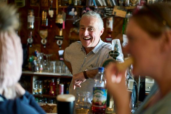Een oudere man, glimlachend en gekleed in een licht shirt, staat achter een bar in een levendige pub in Dublin. Op de voorgrond nipt een vrouw rechts van haar drankje met een rietje, terwijl een ander persoon links wazig is. Flessen en barartikelen vullen de achtergrond en vangen de warme essentie van een onvergetelijke vakantie in Ierland. | Echt Ierland Vakanties