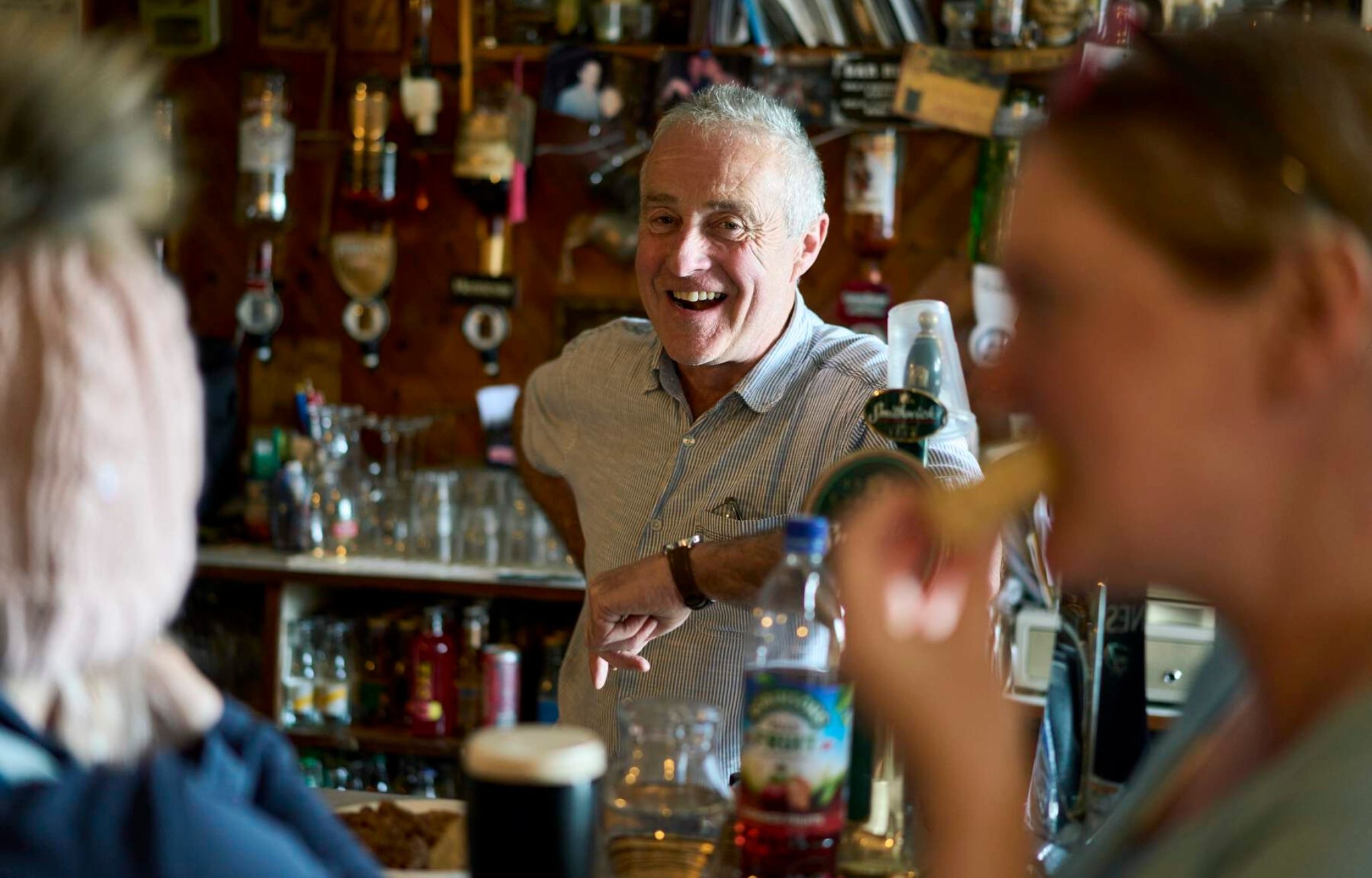 Een oudere man, glimlachend en gekleed in een licht shirt, staat achter een bar in een levendige pub in Dublin. Op de voorgrond nipt een vrouw rechts van haar drankje met een rietje, terwijl een ander persoon links wazig is. Flessen en barartikelen vullen de achtergrond en vangen de warme essentie van een onvergetelijke vakantie in Ierland. | Echt Ierland Vakanties