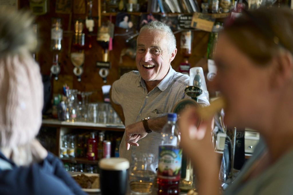 Een man glimlacht warm achter een bar, in gesprek met twee gasten die op de voorgrond zitten. Tegen een achtergrond van verschillende drankjes en decoraties, hint een pint donker bier naar de rijke aanbiedingen die je tegen kunt komen tijdens vakantie in Ierland, en legt de levendige essentie van de scène vast. | Echt Ierland Vakanties
