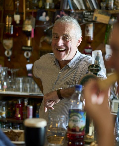Een man glimlacht warm achter een bar, in gesprek met twee gasten die op de voorgrond zitten. Tegen een achtergrond van verschillende drankjes en decoraties, hint een pint donker bier naar de rijke aanbiedingen die je tegen kunt komen tijdens vakantie in Ierland, en legt de levendige essentie van de scène vast. | Echt Ierland Vakanties