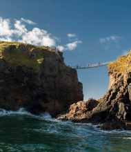 Een touwbrug overspant twee rotsachtige kliffen boven de oceaan en biedt een typische ervaring op elke rondreis Ierland. Vier mensen lopen eroverheen, afgetekend tegen de helderblauwe lucht. De ruige kliffen hebben stukken gras, terwijl golven eronder beuken, met een pad met een leuning zichtbaar op de rechter klif. | Echt Ierland Vakanties
