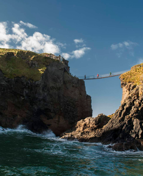 Een touwbrug overspant twee rotsachtige kliffen boven de oceaan en biedt een typische ervaring op elke rondreis Ierland. Vier mensen lopen eroverheen, afgetekend tegen de helderblauwe lucht. De ruige kliffen hebben stukken gras, terwijl golven eronder beuken, met een pad met een leuning zichtbaar op de rechter klif. | Echt Ierland Vakanties