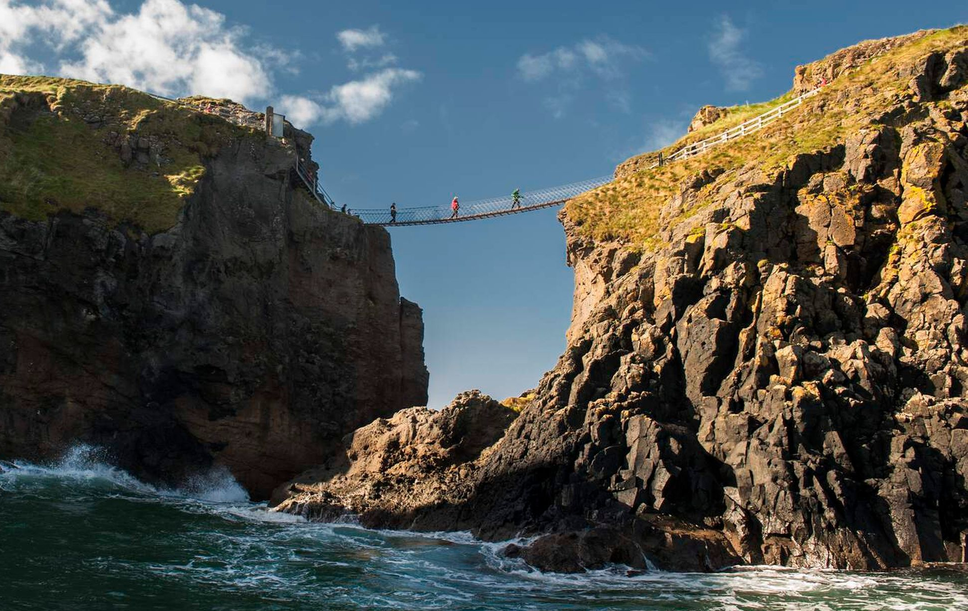 Een touwbrug overspant twee rotsachtige kliffen boven de oceaan en biedt een typische ervaring op elke rondreis Ierland. Vier mensen lopen eroverheen, afgetekend tegen de helderblauwe lucht. De ruige kliffen hebben stukken gras, terwijl golven eronder beuken, met een pad met een leuning zichtbaar op de rechter klif. | Echt Ierland Vakanties