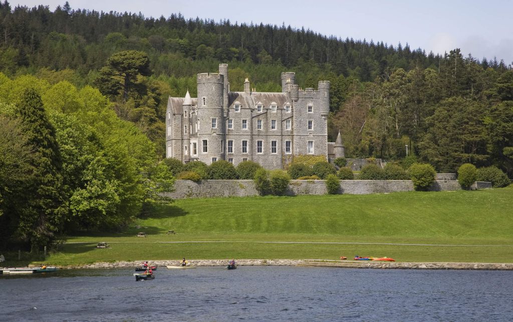 Een groot stenen kasteel met meerdere torentjes staat te midden van weelderig groen, een kenmerk van vakanties in Ierland. Omringd door een dicht bos, kijkt het kasteel uit over een kalm meer, waar mensen in kleine bootjes over het water glijden. De heldere lucht wijst op een perfecte zonnige dag voor een betoverende rundreise Ierland. | Echt Ierland Vakanties