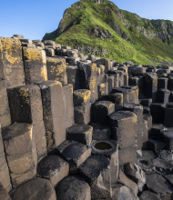 De afbeelding toont de Giant's Causeway in Noord-Ierland, een must-see op elke rondreis Ierland. Grote, in elkaar grijpende hexagonale basaltkolommen domineren de voorgrond, terwijl een groene, rotsachtige heuvel oprijst tegen een helderblauwe lucht op de achtergrond. Gele korstmos stipt sommige van de kolommen. | Echt Ierland Vakanties