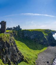 Panoramisch uitzicht op een oud stenen kasteel op een met gras begroeide klif met uitzicht op de oceaan: een perfect uitzicht voor uw Urlaub Irland. De ruïnes, met boogramen, worden omarmd door weelderig groen landschap. Beneden beuken golven op een rotsachtige kustlijn onder de helderblauwe lucht terwijl de horizon zich uitstrekt tot in de verte. | Echt Ierland Vakanties
