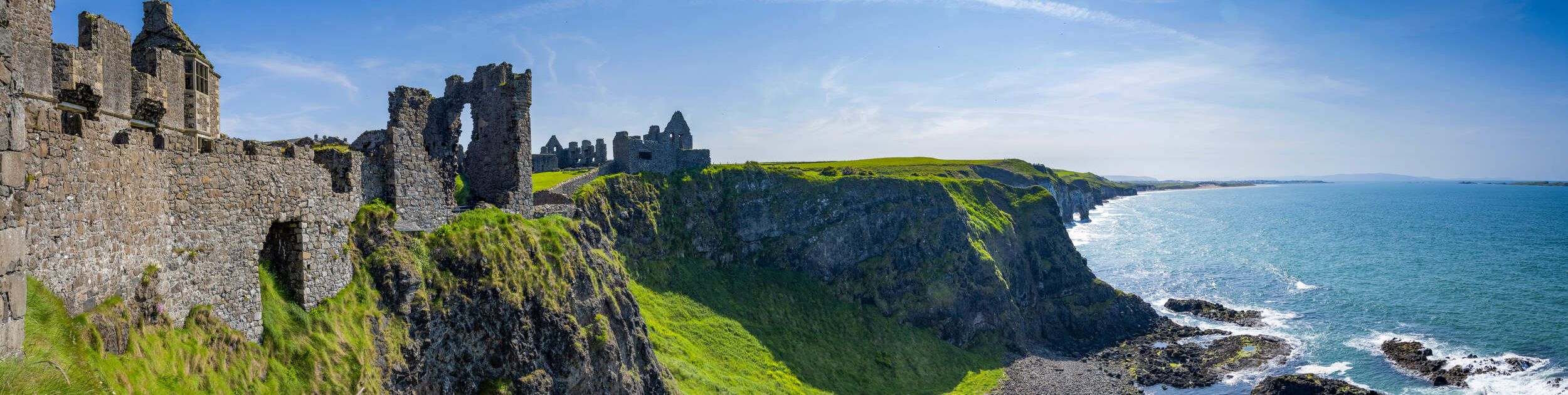 Panoramisch uitzicht op een oud stenen kasteel op een met gras begroeide klif met uitzicht op de oceaan: een perfect uitzicht voor uw Urlaub Irland. De ruïnes, met boogramen, worden omarmd door weelderig groen landschap. Beneden beuken golven op een rotsachtige kustlijn onder de helderblauwe lucht terwijl de horizon zich uitstrekt tot in de verte. | Echt Ierland Vakanties