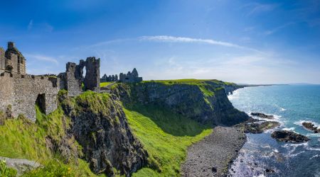 Panoramisch uitzicht op een oud stenen kasteel op een met gras begroeide klif met uitzicht op de oceaan: een perfect uitzicht voor uw Urlaub Irland. De ruïnes, met boogramen, worden omarmd door weelderig groen landschap. Beneden beuken golven op een rotsachtige kustlijn onder de helderblauwe lucht terwijl de horizon zich uitstrekt tot in de verte. | Echt Ierland Vakanties