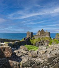 Ontdek het betoverende uitzicht op de ruïnes van Dunluce Castle op een ruige klif met uitzicht op de zee, perfect voor uw rundreise Ierland. Stenen muren omlijsten dit historische bouwwerk, omgeven door weelderig groen gras tegen een achtergrond van gedeeltelijk bewolkte luchten en blauwe vlekken. | Echt Ierland Vakanties