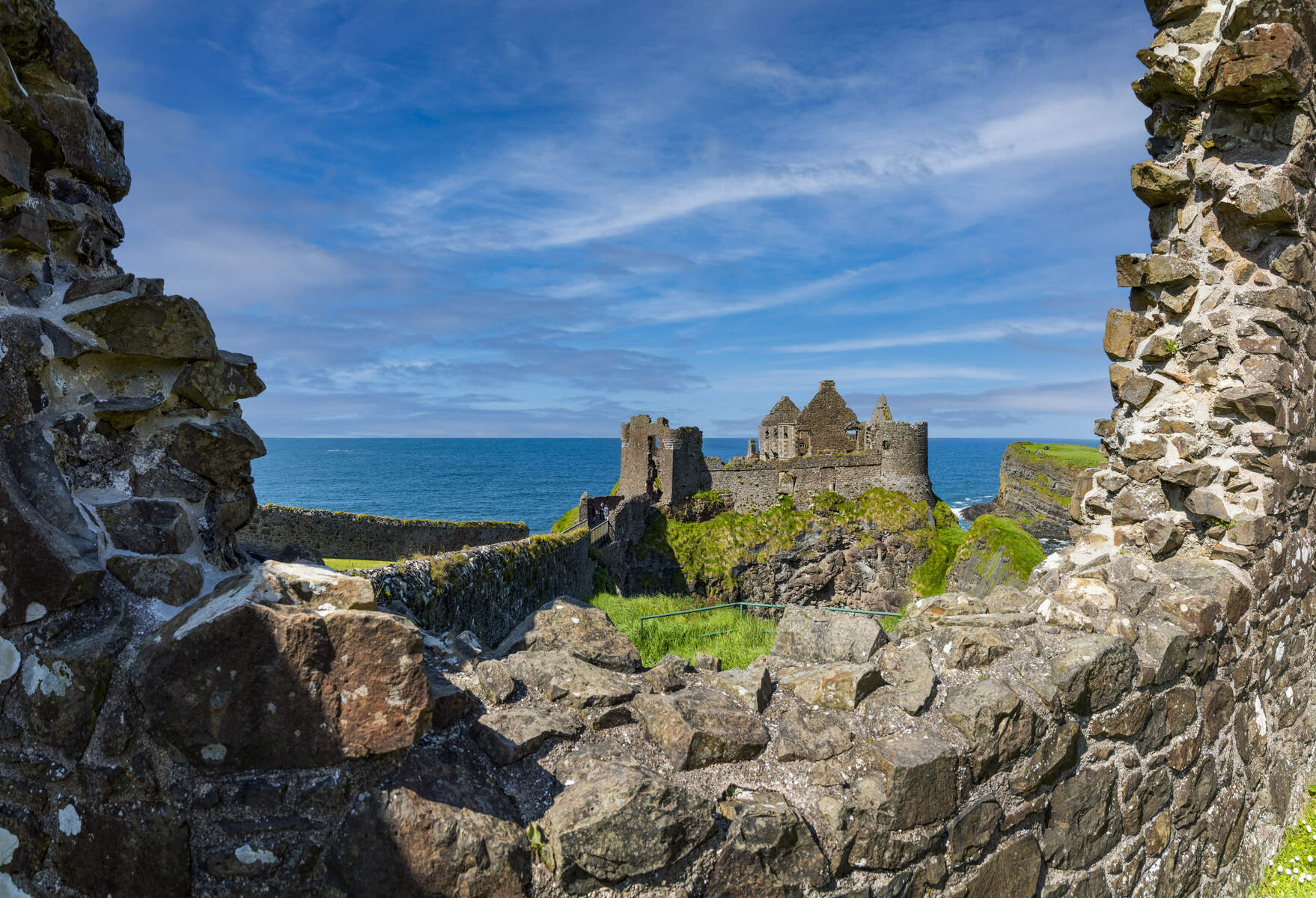 Ontdek het betoverende uitzicht op de ruïnes van Dunluce Castle op een ruige klif met uitzicht op de zee, perfect voor uw rundreise Ierland. Stenen muren omlijsten dit historische bouwwerk, omgeven door weelderig groen gras tegen een achtergrond van gedeeltelijk bewolkte luchten en blauwe vlekken. | Echt Ierland Vakanties