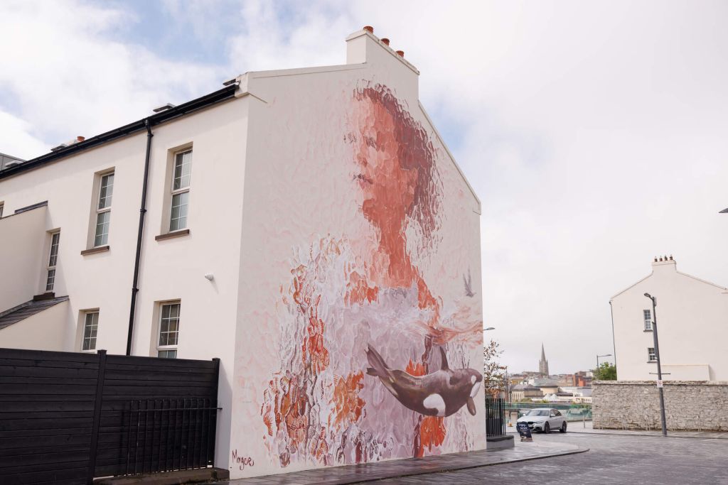 Een grote muurschildering op de zijkant van een wit gebouw in Ierland toont een vrouw met wazige trekken die een orka vasthoudt. De kunst combineert zachte roze en oranje tinten. De straat is nat, wat duidt op recente regenval, en een kerktoren is zichtbaar op de achtergrond onder een bewolkte lucht. | Echt Ierland Vakanties