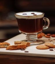 Een glazen mok met koffie, gegarneerd met slagroom en koffiebonen, staat op een houten oppervlak en roept de warmte van een vakantie in Ierland op. In de buurt liggen diverse koekjes, steranijs, kaneelstokjes en verspreide koffiebonen op een witte snijplank. De zacht wazige achtergrond versterkt de gezellige ambiance. | Echt Ierland Vakanties