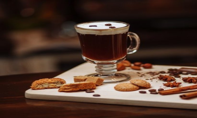 Een glazen mok met koffie, gegarneerd met slagroom en koffiebonen, staat op een houten oppervlak en roept de warmte van een vakantie in Ierland op. In de buurt liggen diverse koekjes, steranijs, kaneelstokjes en verspreide koffiebonen op een witte snijplank. De zacht wazige achtergrond versterkt de gezellige ambiance. | Echt Ierland Vakanties