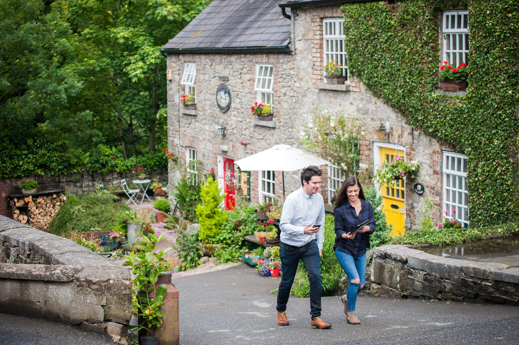 Een man en een vrouw slenteren over een pad langs een stenen huis versierd met klimop en bloemen, wat doet denken aan betoverende vakanties in Ierland. De man in een lichtblauw shirt en donkere broek houdt een telefoon vast, terwijl de vrouw in donkere kleding hetzelfde doet. Een felgele deur en tuinmeubilair maken dit idyllische tafereel compleet. | Echt Ierland Vakanties