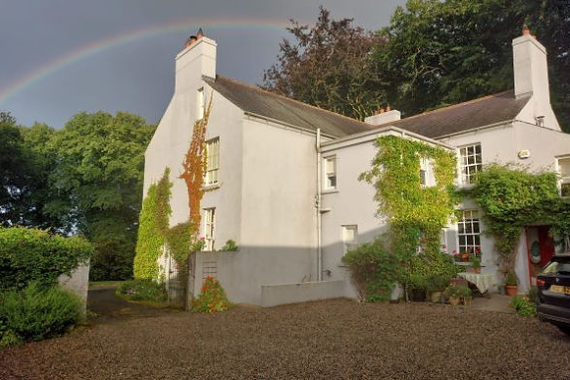Een schilderachtig, lichtgrijs huis met witte schoorstenen is versierd met klimop, wat doet denken aan charmante Ierse huisjes die perfect zijn voor Reisen Irland. Tegen de achtergrond van donkere bomen onder een gedeeltelijk bewolkte lucht, een levendige regenboog die erboven bogen. De grindoprit is omzoomd met potplanten bij de ingang. | Echt Ierland Vakanties