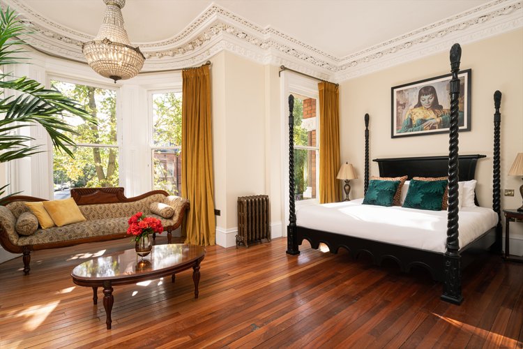 Elegante slaapkamer die doet denken aan charmante Ierse interieurs, met een donkerhouten hemelbed versierd met blauwgroene en bruine kussens. Een vintage bank bij grote ramen met gele gordijnen roept een "Ierland vakanties"-sfeer op, terwijl de hardhouten vloer de sierlijke kroonluchter en decoratieve plafondlijsten aanvult. | Echt Ierland Vakanties