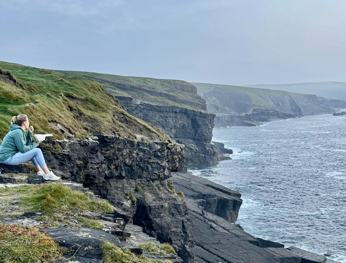 Een persoon in een groen jasje en jeans zit op een rotsachtige klif, perfect vastgelegd voor een vakantiebrochure over Ierland. Ze houden een notitieboekje vast en schetsen de ruige kustlijn. De zee strekt zich uit tot aan de mistige horizon onder een bewolkte lucht, terwijl weelderig groen gras contrasteert met de donkere, grillige rotsen eronder. | Echt Ierland Vakanties