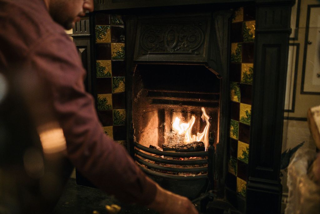 Iemand in een rood overhemd staat bij de brandende open haard met een sierlijk zwart metalen frame en decoratieve geelgroene tegels, waardoor er een warme en gezellige sfeer binnenshuis ontstaat. | Echt Ierland Vakanties
