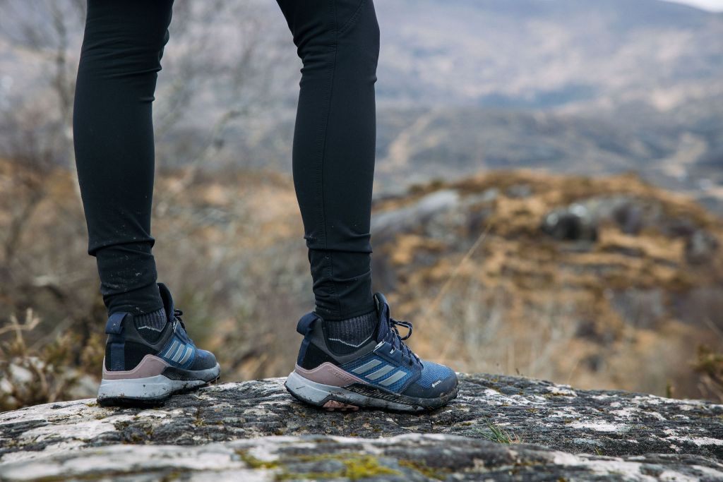 Een persoon in een donkere legging en blauwe trailschoenen staat op een rotsachtige ondergrond en kijkt uit over een wazig berglandschap met stukken bruin gras en rotsen op de achtergrond. De scène suggereert een wandeling of een buitenavontuur. | Echt Ierland Vakanties